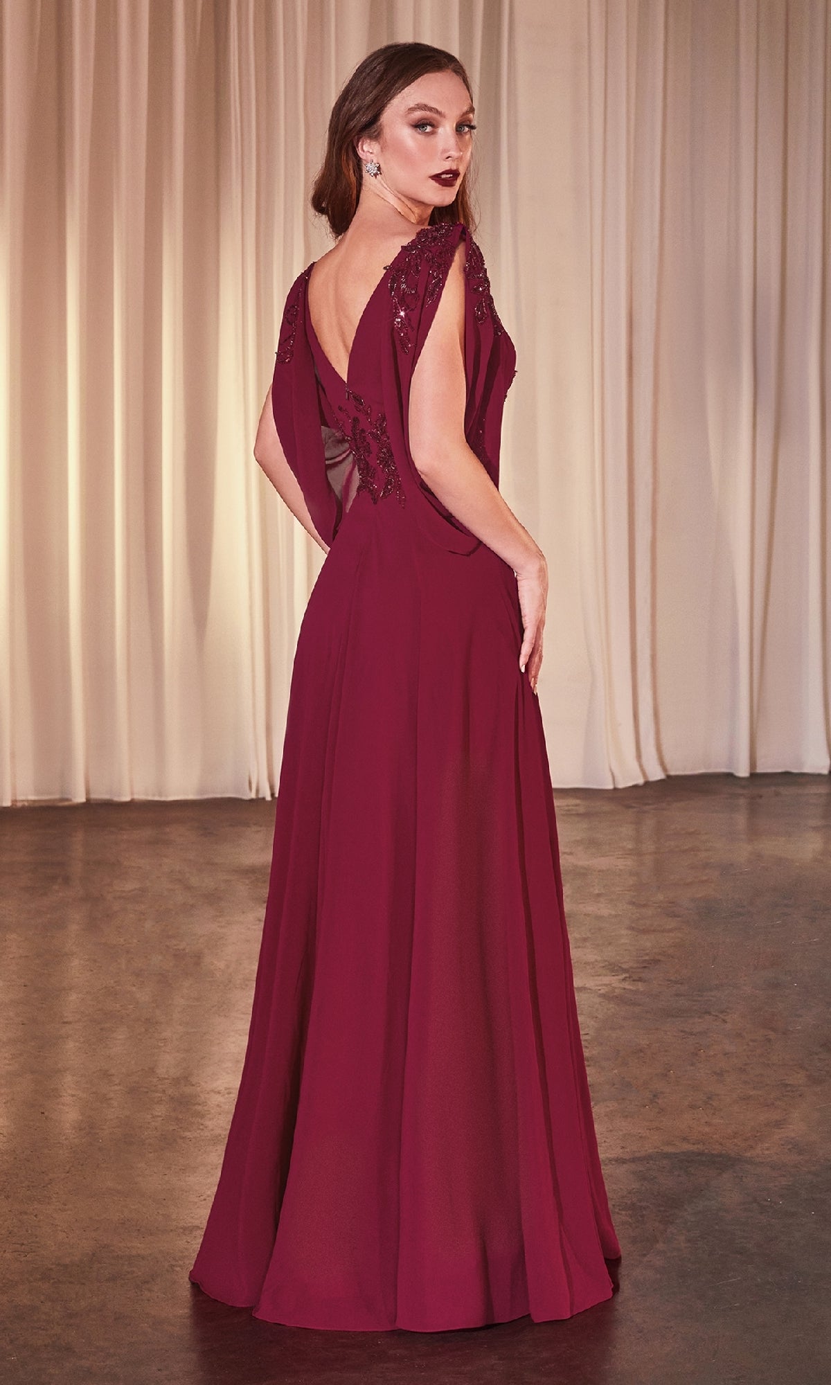 A woman wears the Ladivine CDS508 Cowl-Sleeve Long Formal Dress, featuring deep red fabric and back embroidery, as she stands on a polished floor with cream curtains behind her, facing away from the camera.